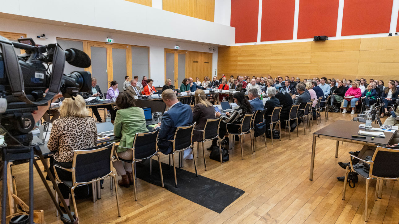 Sitzung eines Ausschusses an mehreren rechteckig angeordneten Tischen mit Medienbeteiligung und einer Kamera links im Bild sowie Publikum vor einer Wand 